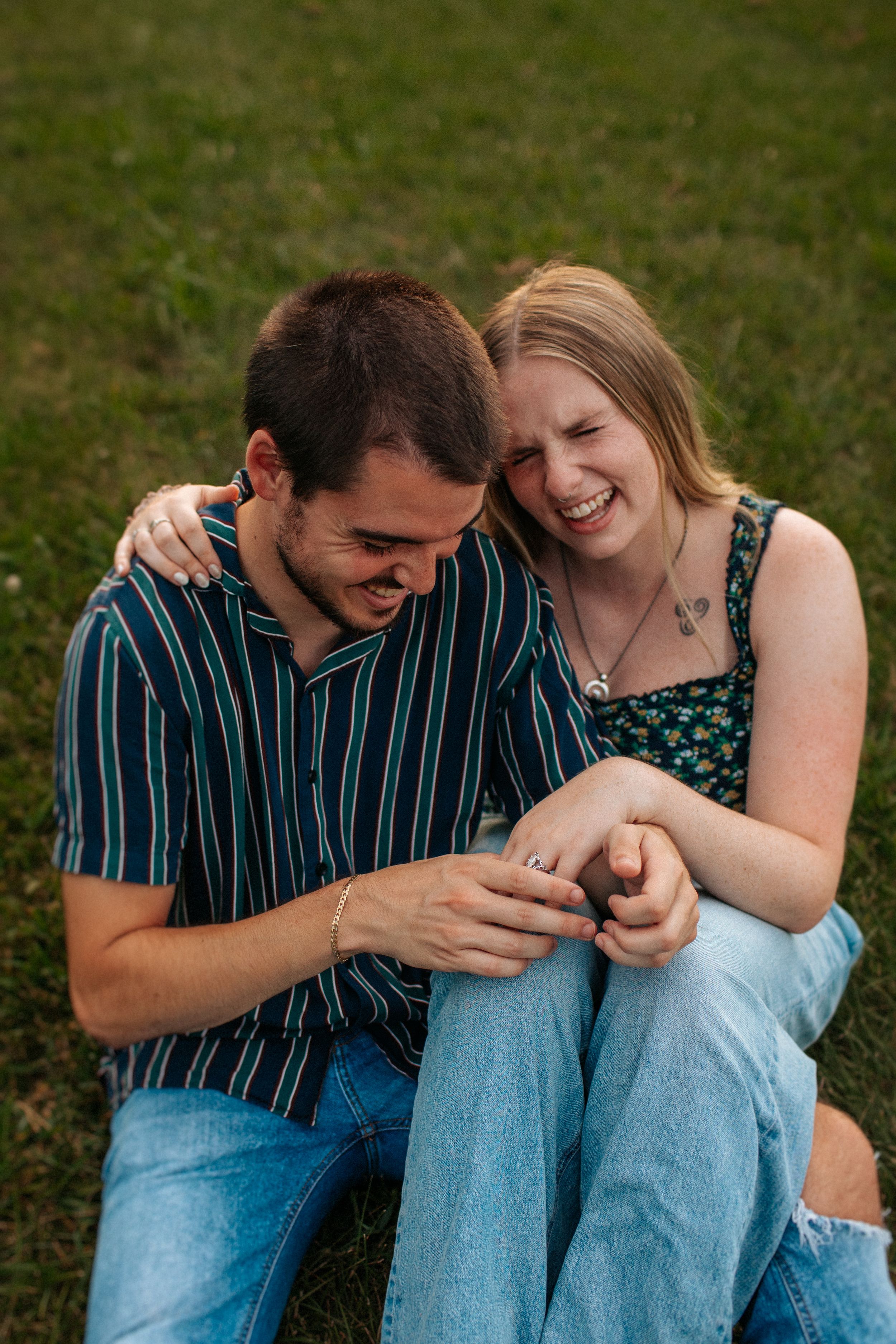 A couple sits close together on grass, sharing laughter while looking at something on the ground.