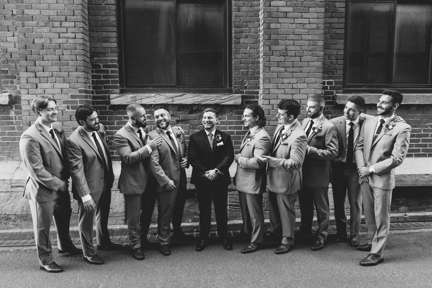 A vintage black and white photograph shows a row of people sitting on a brick ledge against a building wall.
