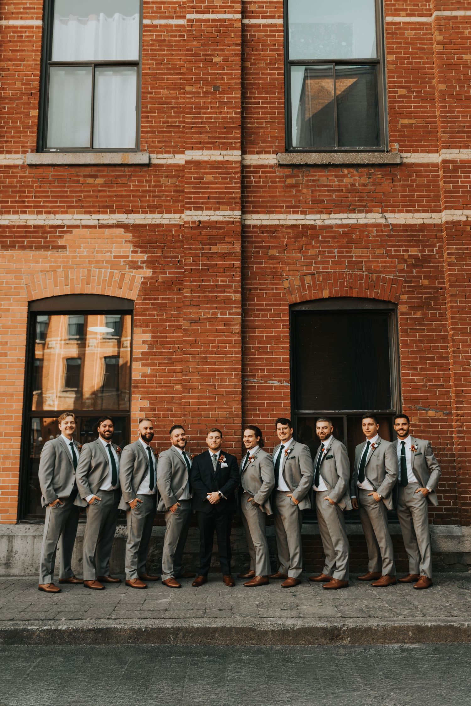 A wedding party in matching gray suits sits casually along a brick wall for a creative group photo.