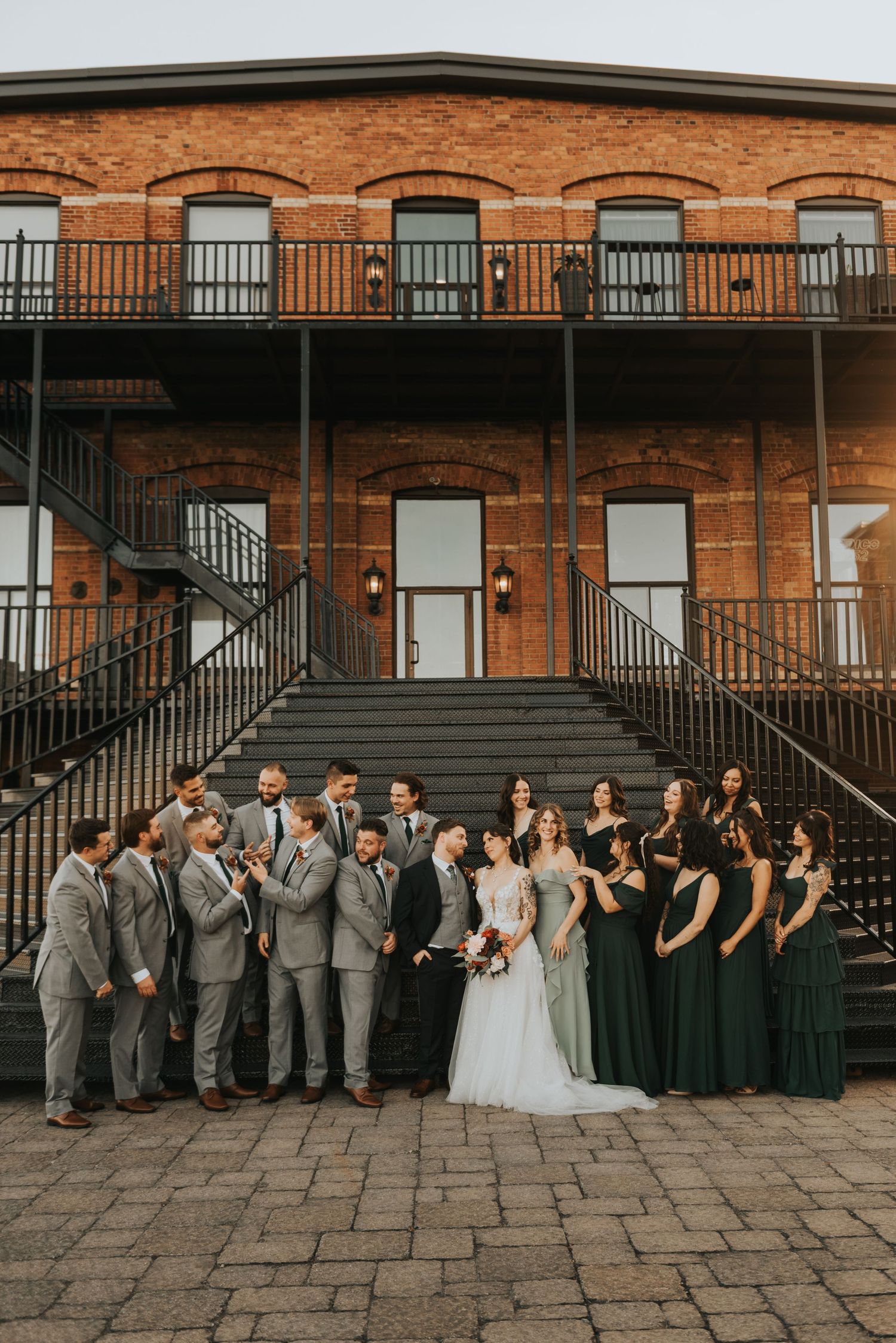 Wedding party poses on brick steps wearing gray suits and black dresses at historic building exterior.