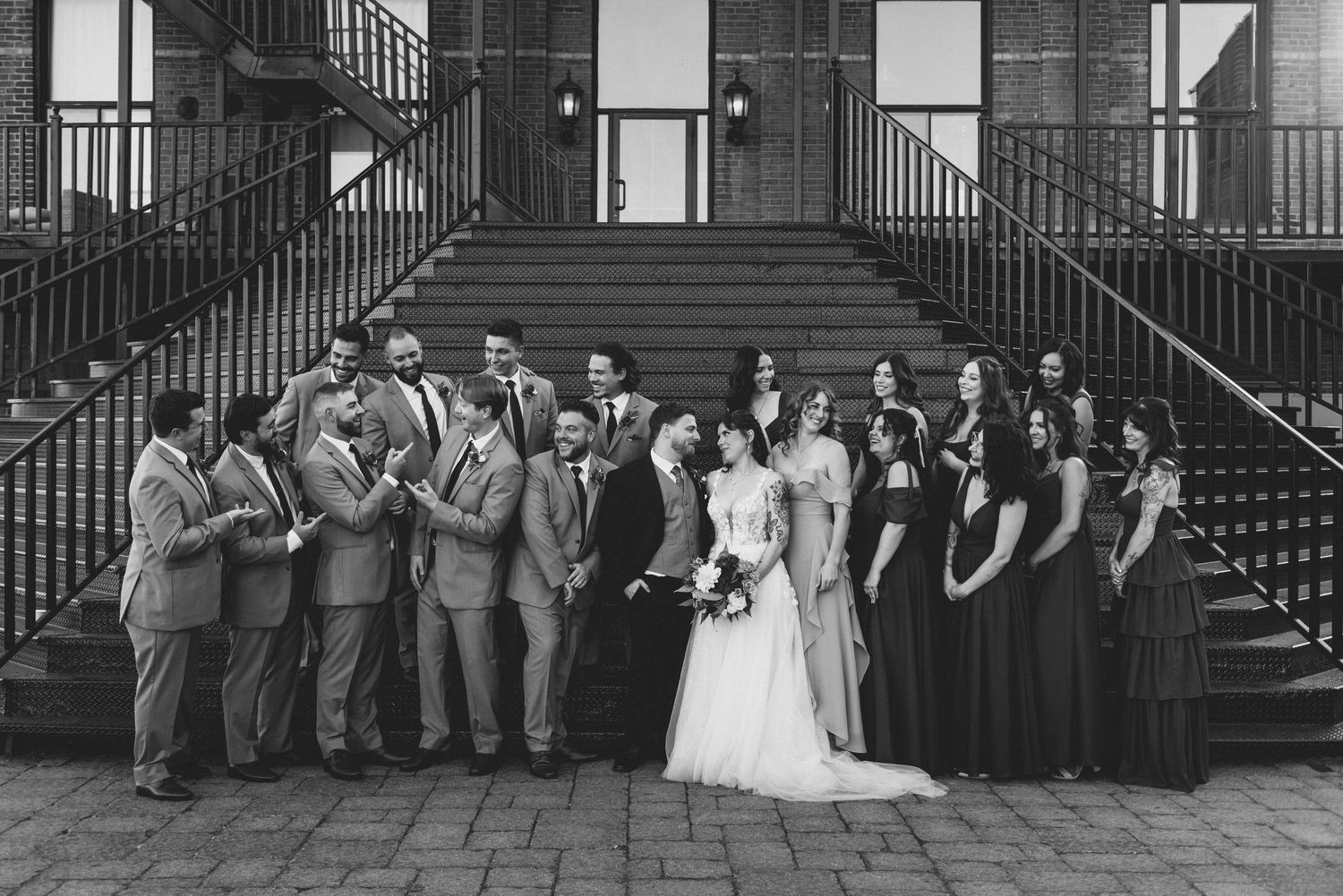 A black and white wedding party photo with groomsmen in suits and bridesmaids in dark dresses posed on grand stone steps.