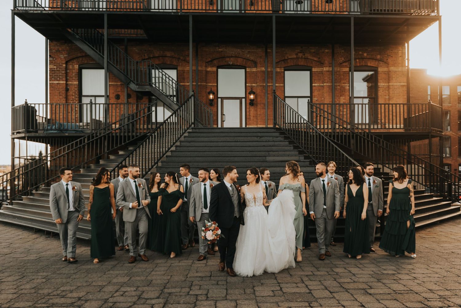 Wedding party members stand in a line against a rustic brick building during golden hour.