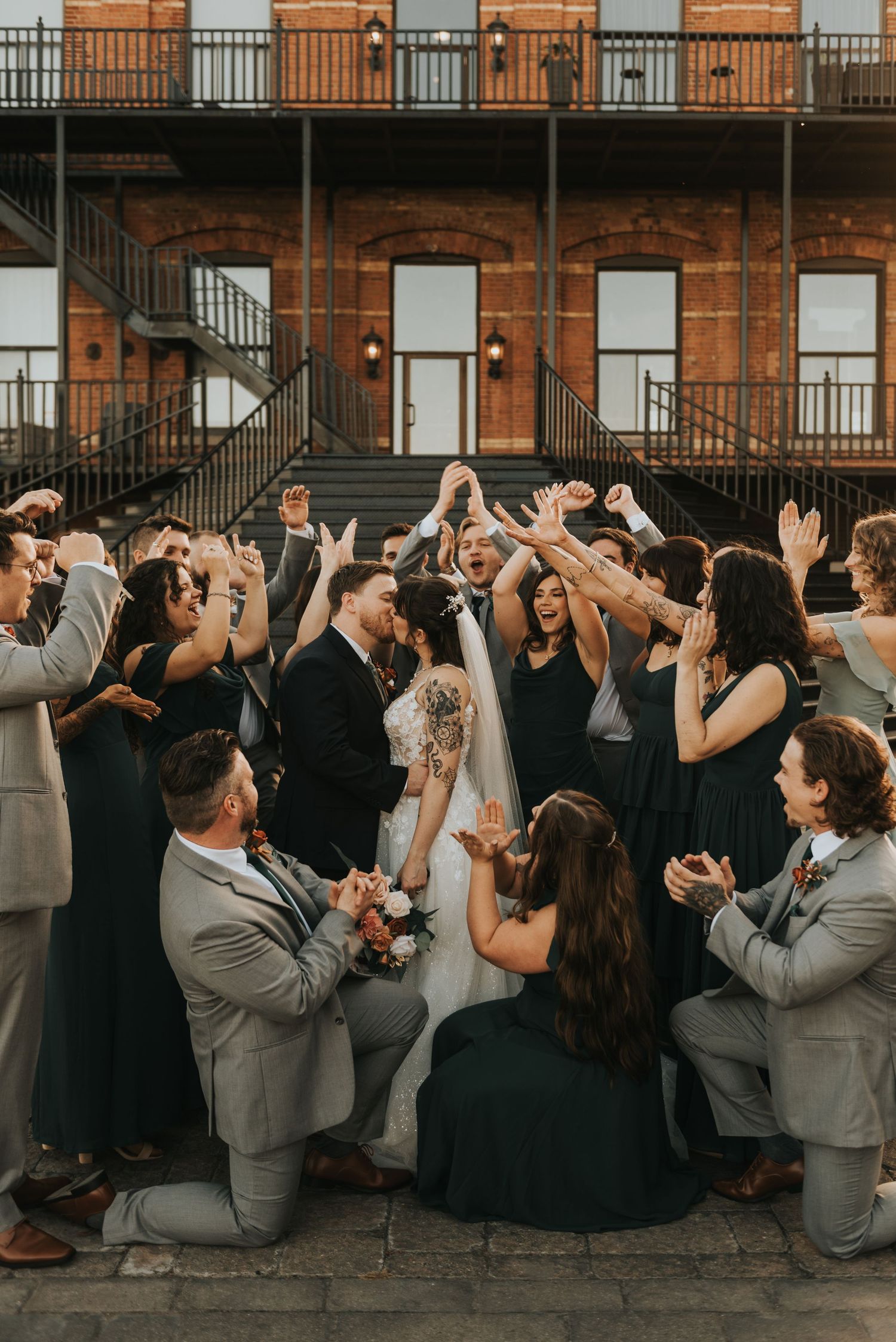Wedding party celebrates with raised arms and cheers against brick building backdrop in formal attire.