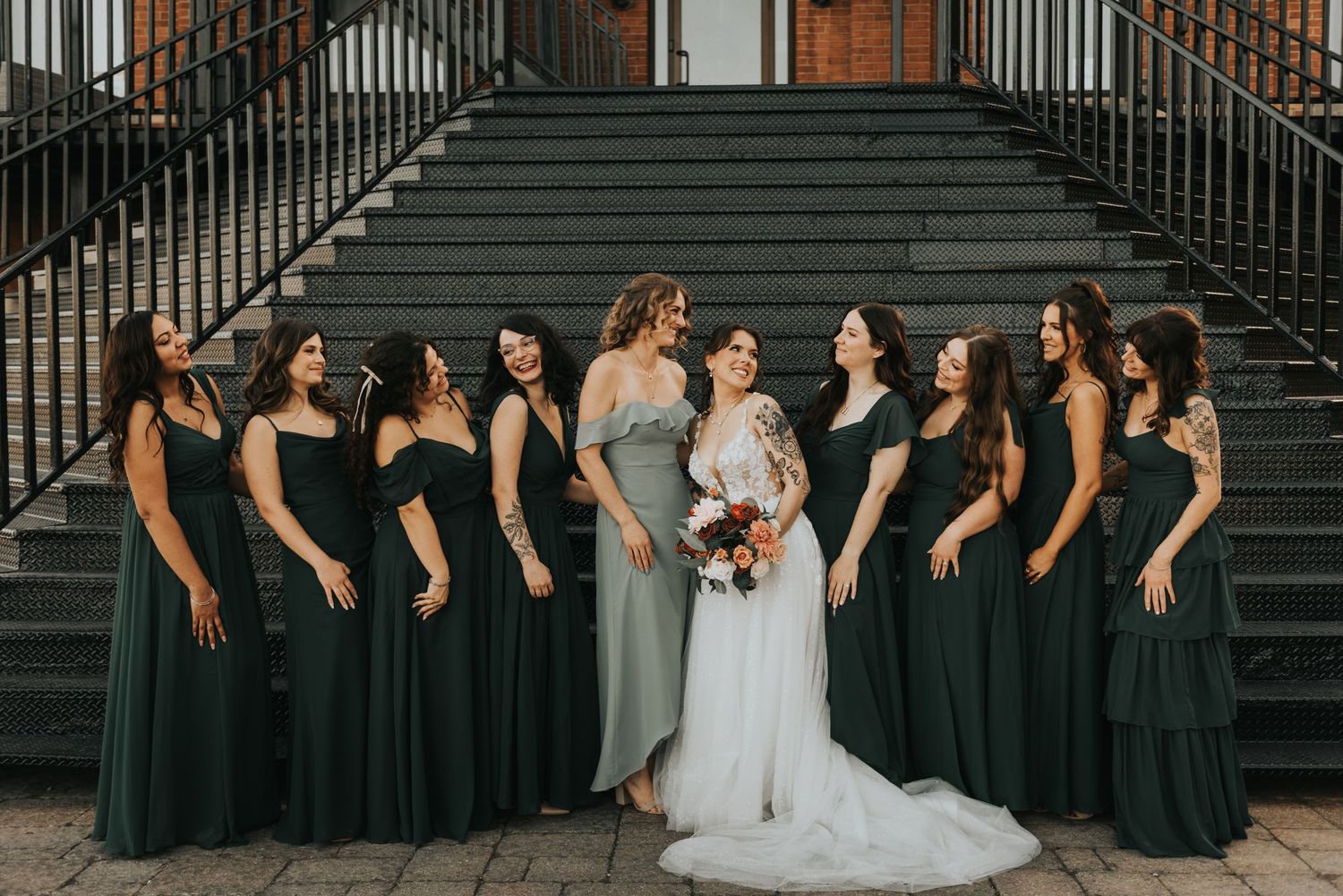 Wedding party wearing dark green bridesmaid dresses and a white bridal gown pose on elegant brick staircase.