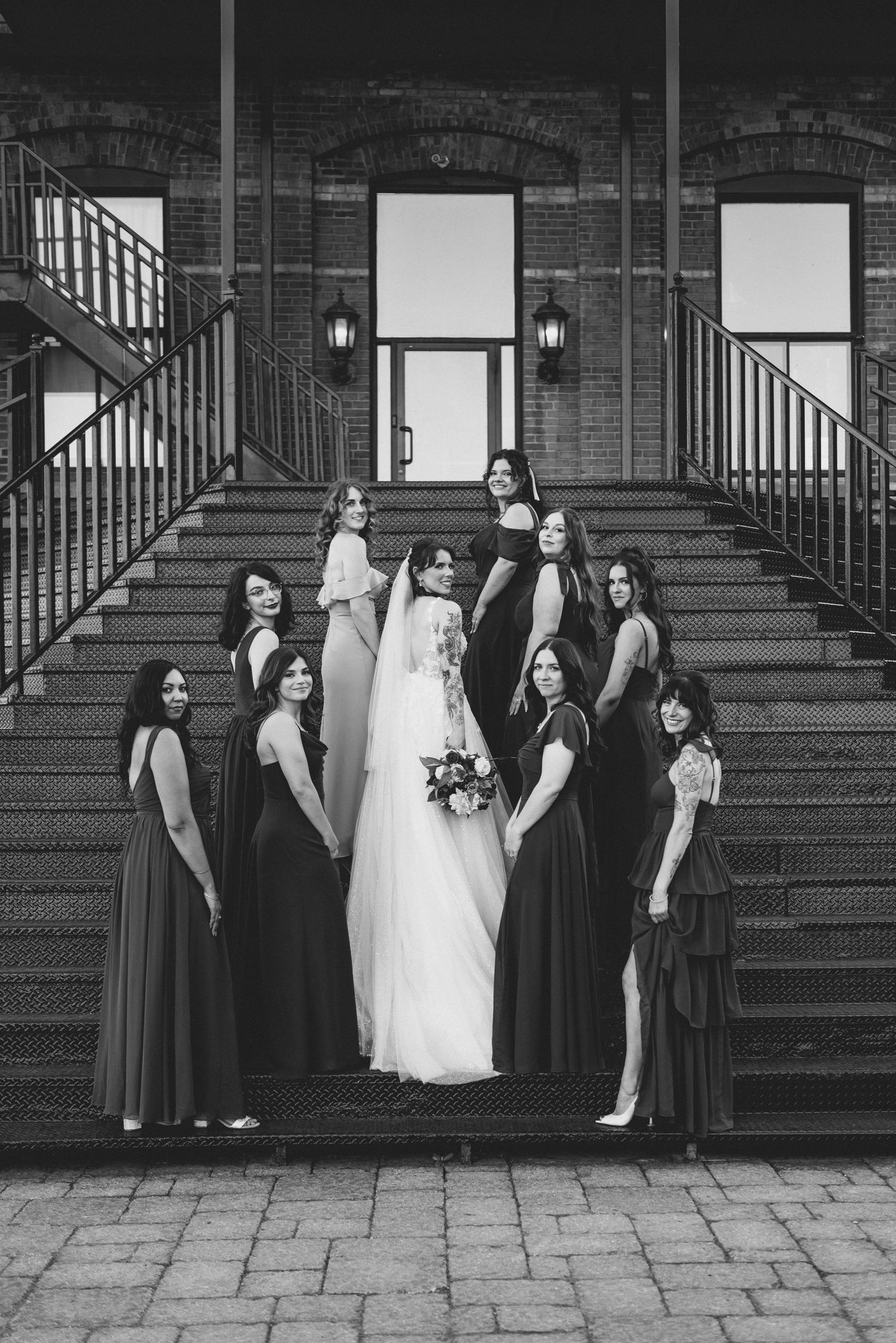 Black and white photo of wedding party arranged on brick staircase against industrial building backdrop.