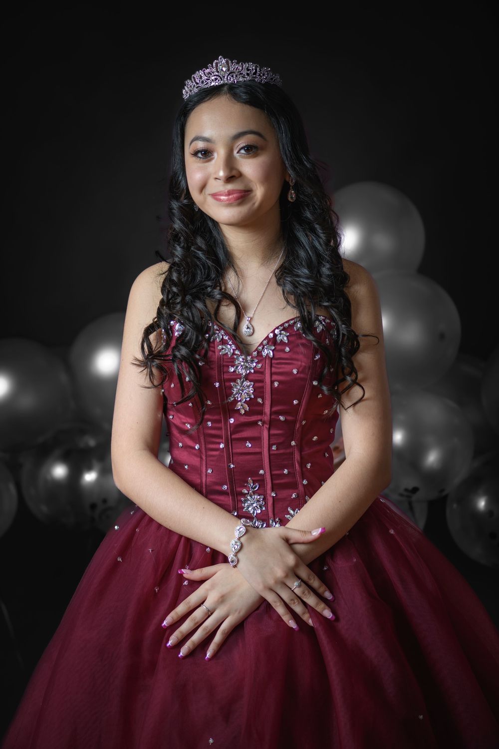 Teenage girl in burgundy quinceañera dress and tiara smiling with balloons in the background.