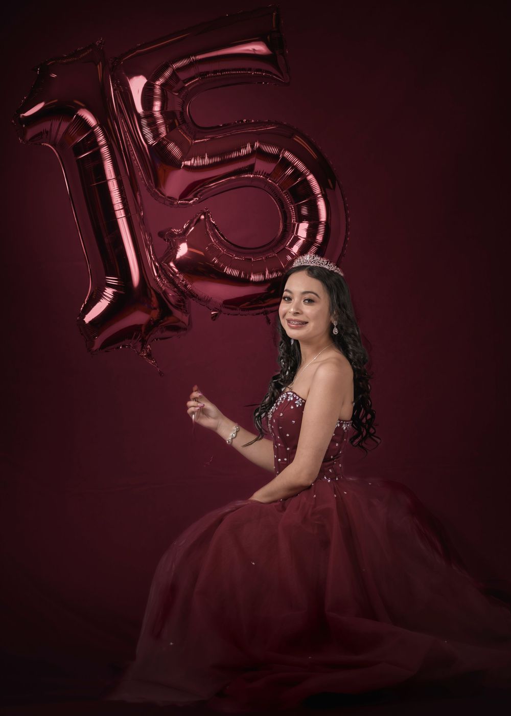 Young girl in burgundy quinceañera gown posing with number 15 balloons on dark red background.