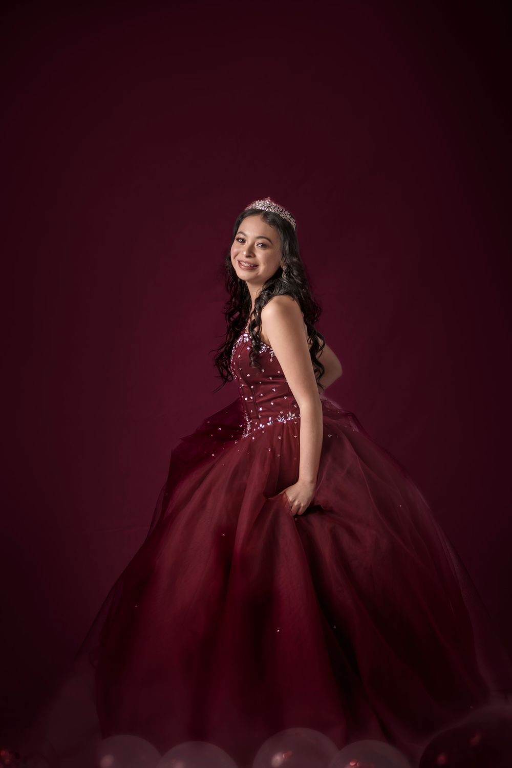 Young woman in elegant burgundy ball gown and tiara posing against a dark red background.