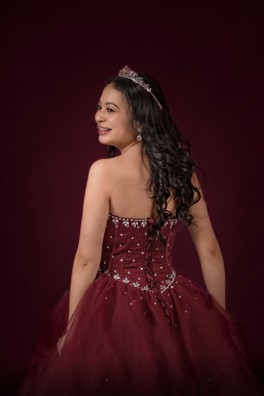 Back view of a young woman in a strapless burgundy quinceañera gown with jeweled details and tiara.