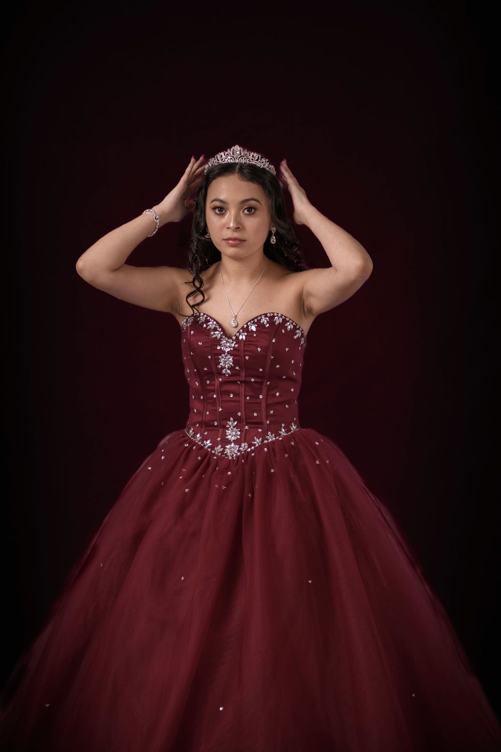 Studio photograph of a young woman wearing a red ball gown and a tiara with her hands up holding her crown