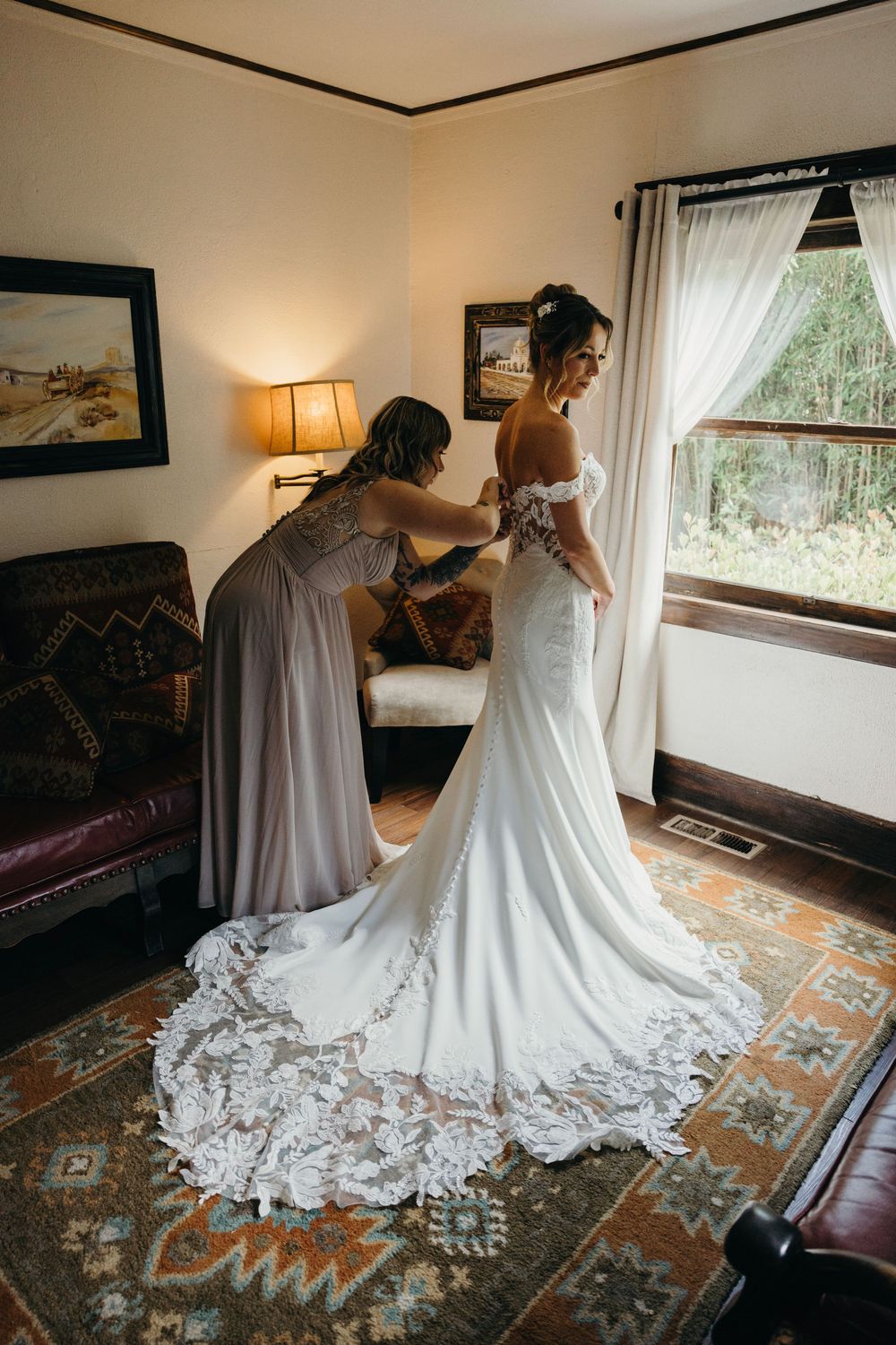 Wedding dress with intricate lace train spreads across patterned carpet during preparation.
