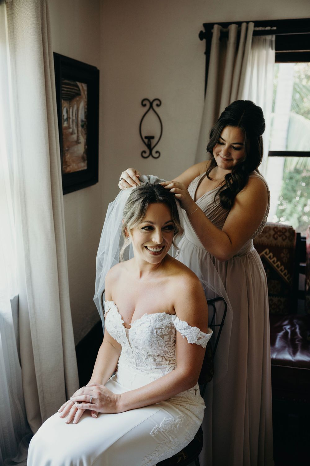 A bride in a strapless lace wedding gown smiles while getting ready in a sunlit room.