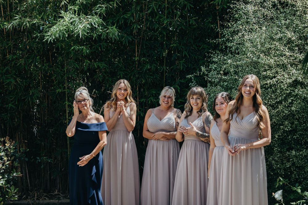 Bridesmaids in light neutral dresses stand together outdoors surrounded by greenery.