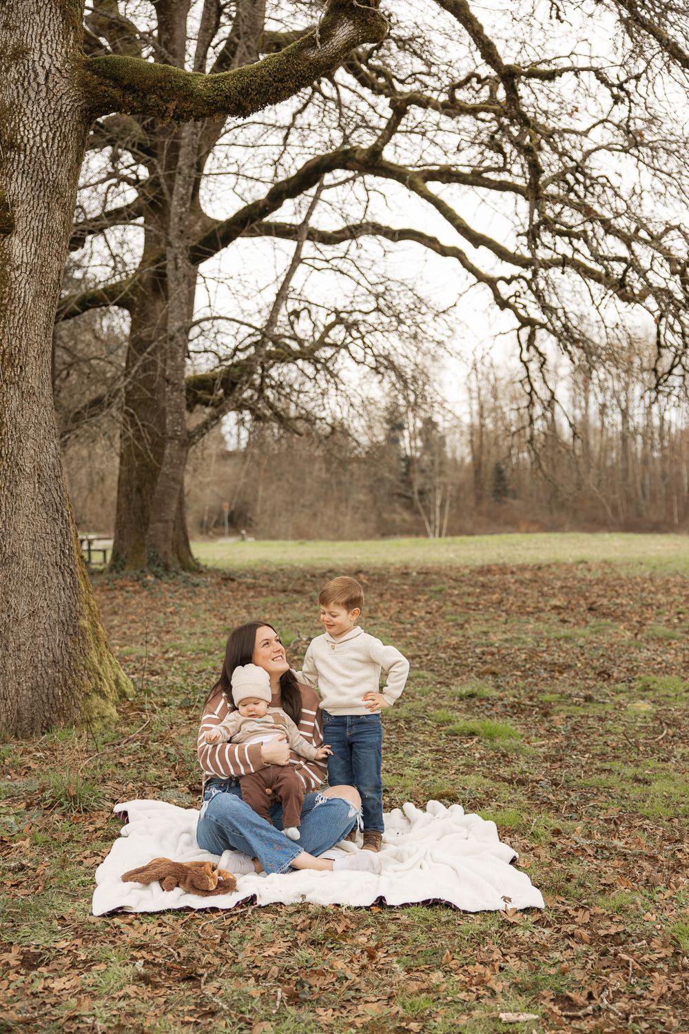 A mother sits on a white blanket with her two young children under a large bare oak tree in a park.