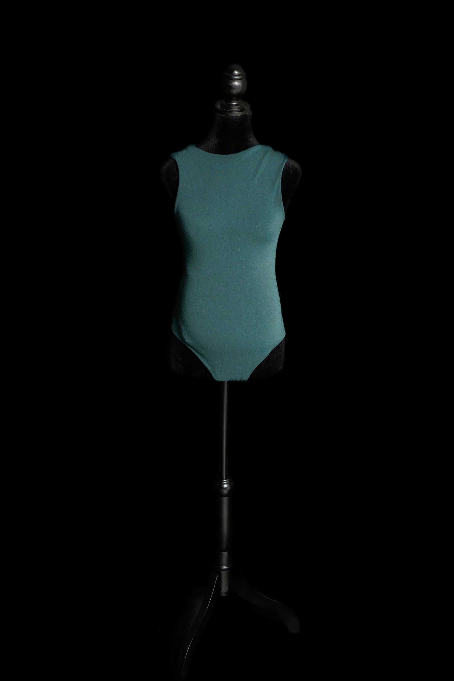 Teal sleeveless maternity bodysuit with high neckline and fitted silhouette displayed on a dress form.