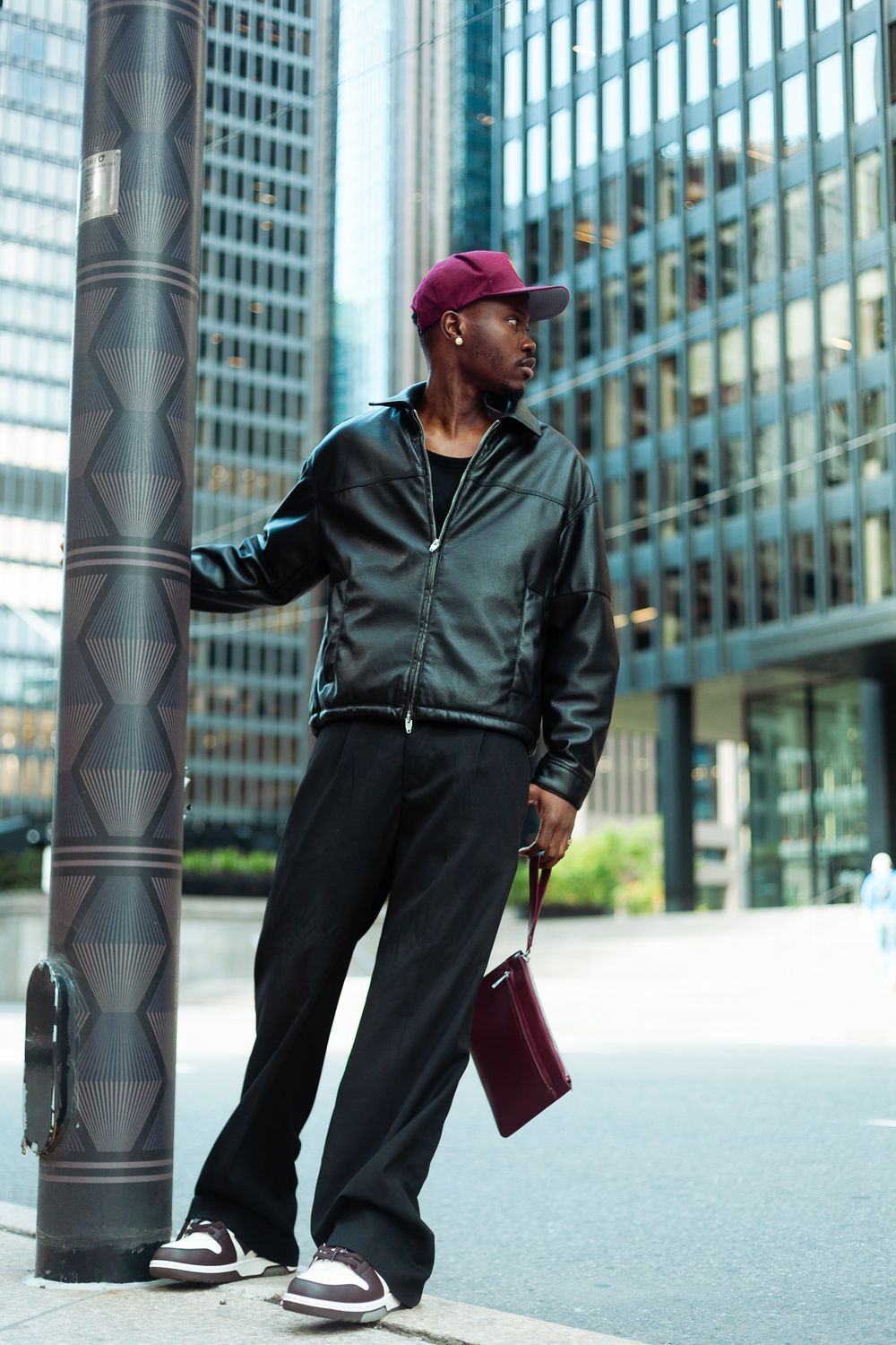 Stylish man in black leather jacket and burgundy cap leaning on urban pole.