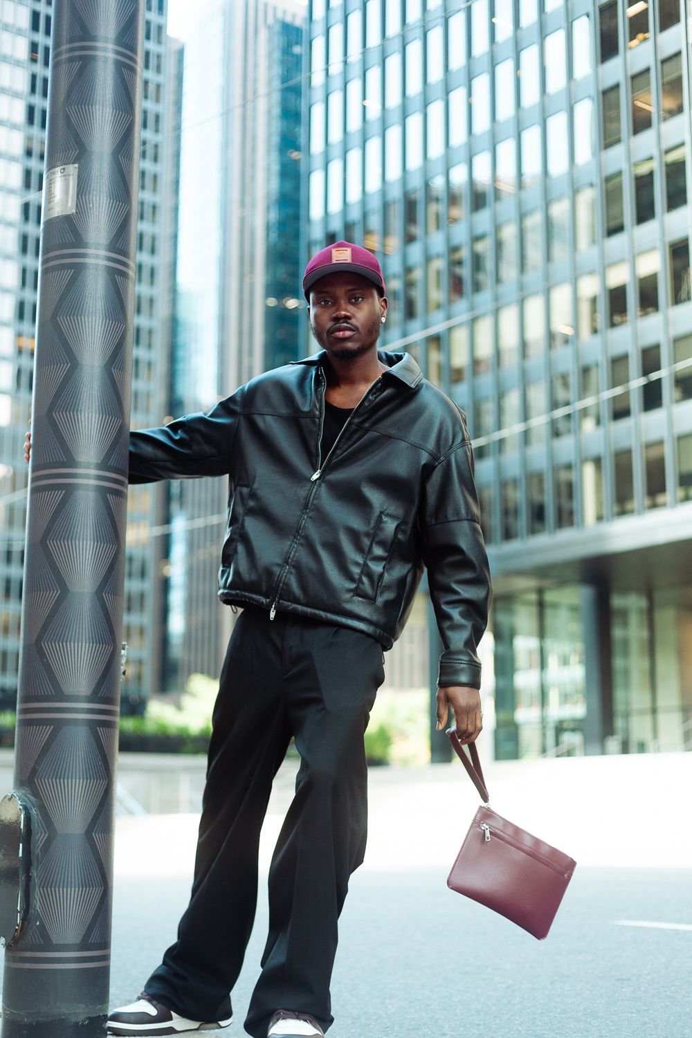 Fashionable man holding burgundy clutch bag leaning on pole in city setting.