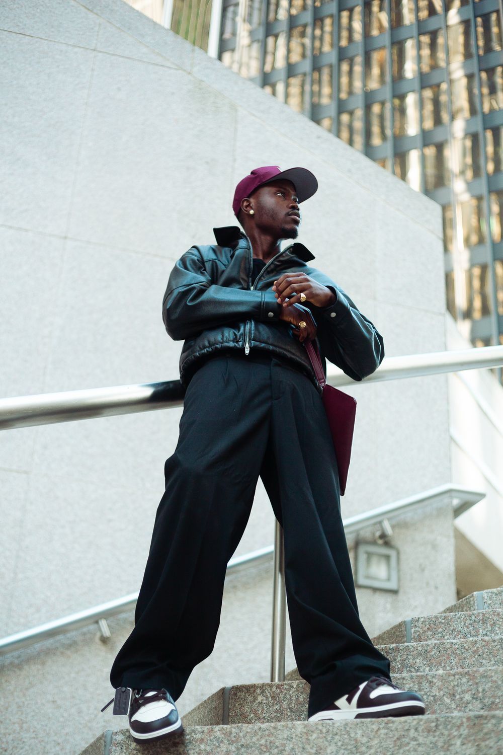 Young Black man in black leather jacket, wide-leg pants, burgundy cap, and Jordan sneakers posing on urban steps.