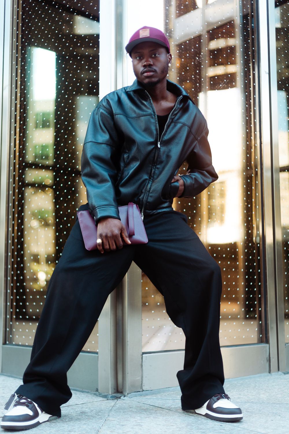 Stylish Black man in leather jacket, wide-leg black pants, and burgundy cap holding a folder against a glass door.