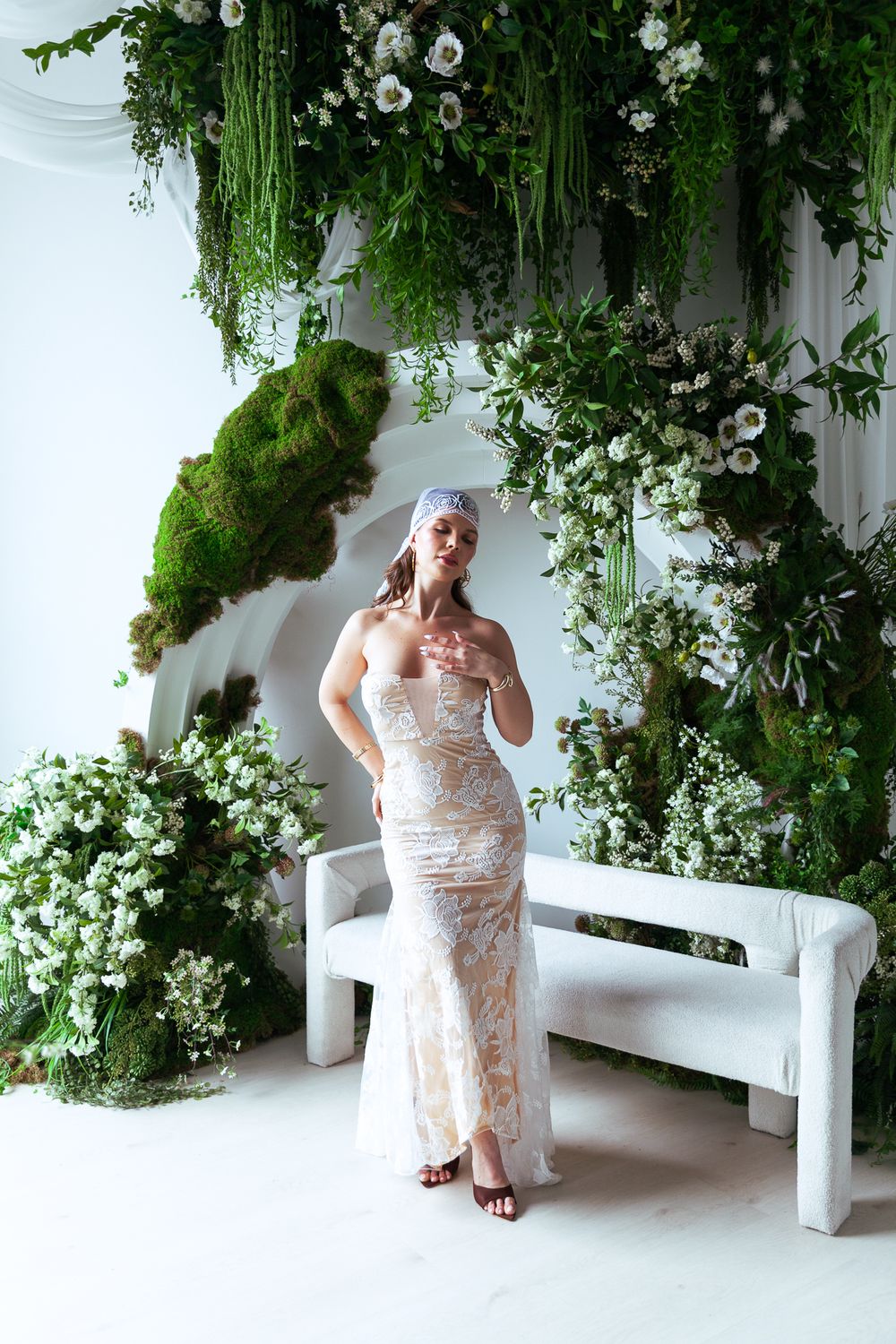 Bride in lace gown with blue cap veil standing under lush floral and greenery installation.