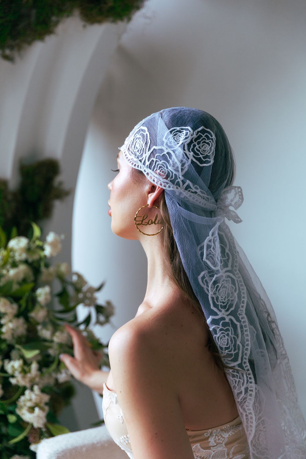 Bride in profile wearing blue lace veil headpiece with rose embroidery near white floral arch.