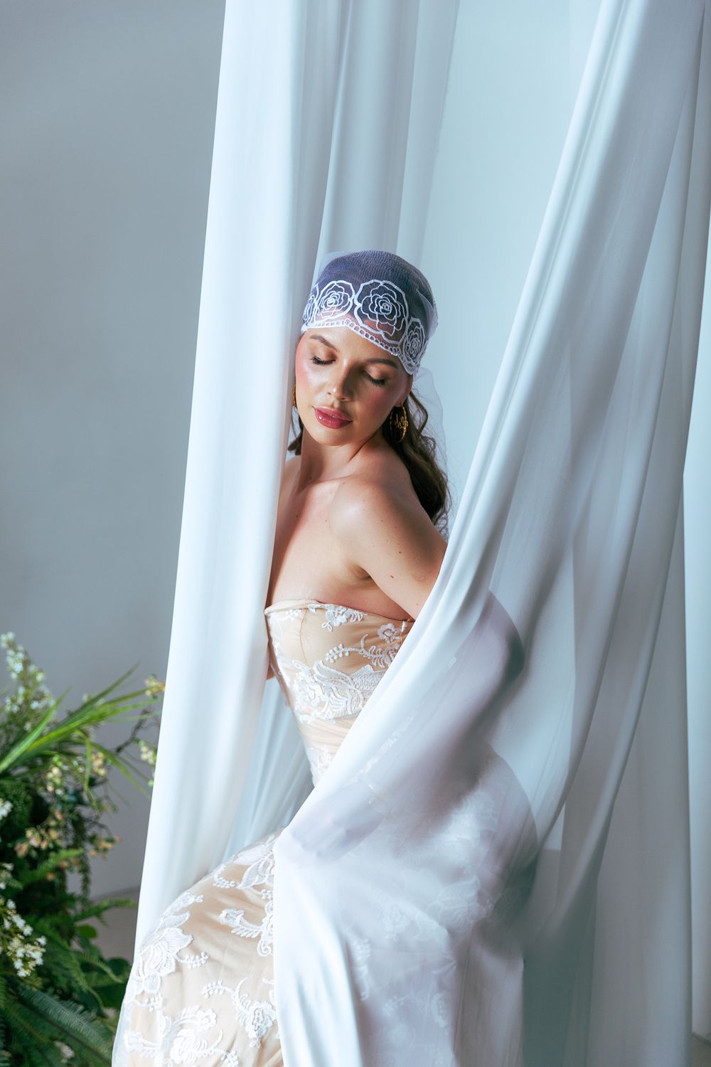 Bride in strapless lace wedding gown and blue embroidered headpiece poses between flowing white curtains.