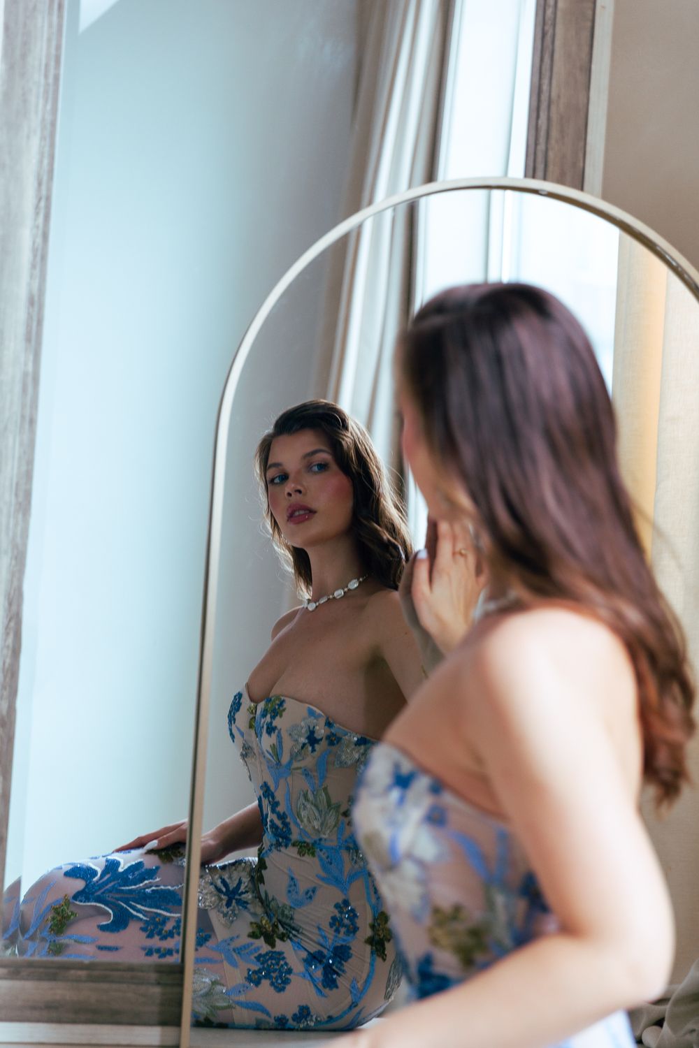 Brunette woman in floral strapless blue dress gazes at her reflection in an arched floor mirror.