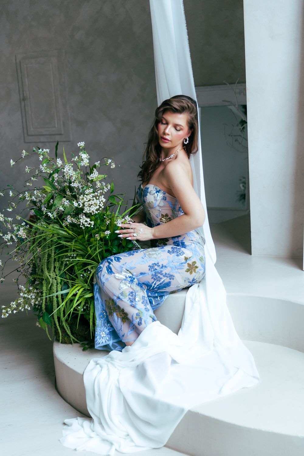 Woman in strapless blue floral gown sits beside lush white wildflowers with draped white fabric.