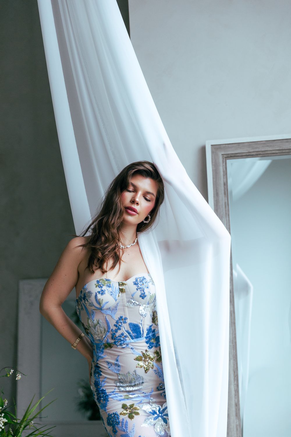 Woman in strapless blue floral dress with pearl necklace poses beside flowing white fabric near mirror.