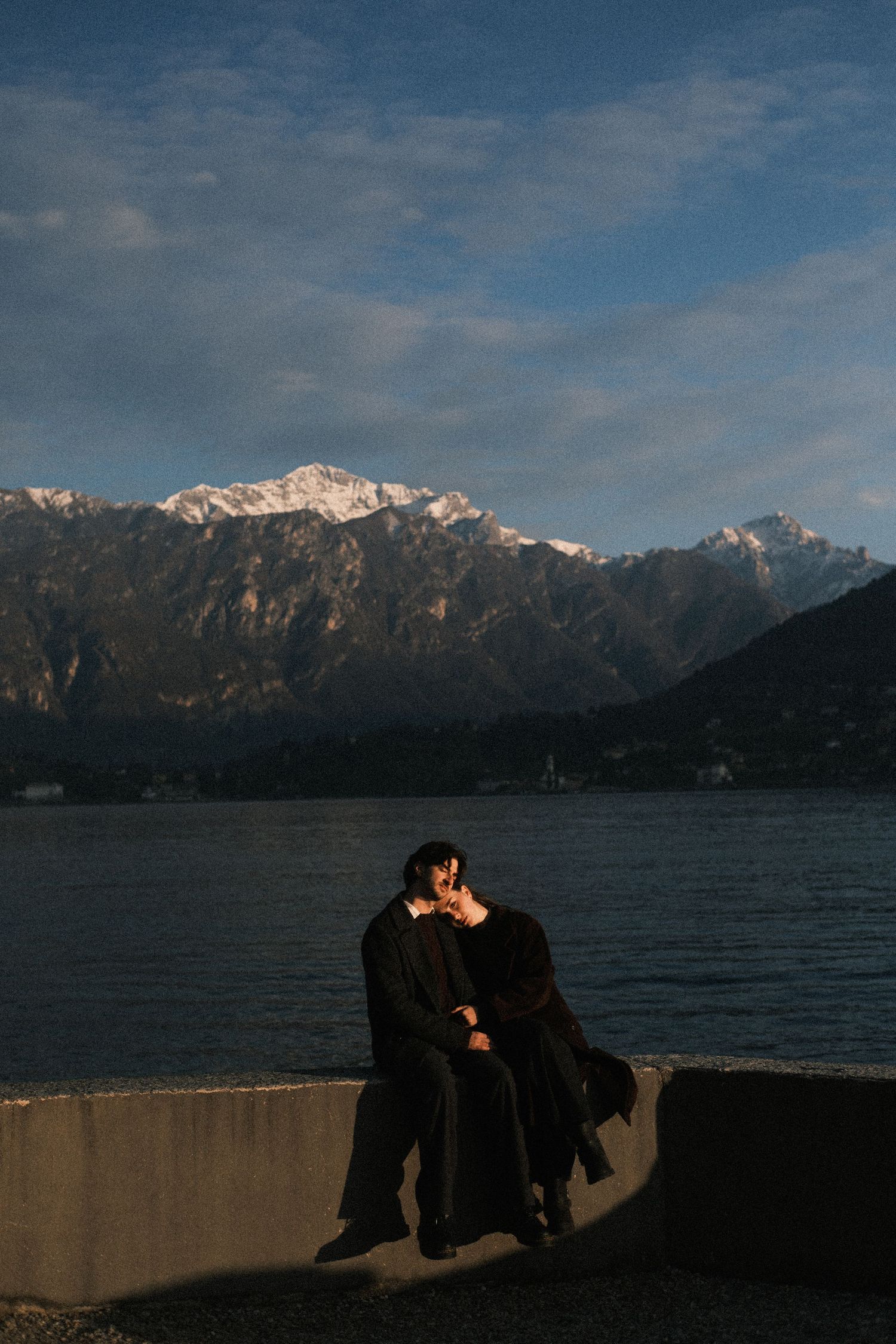 Silhouetted couple embraces on seawall with snow-capped mountains and moody blue sky in backdrop at sunset.
