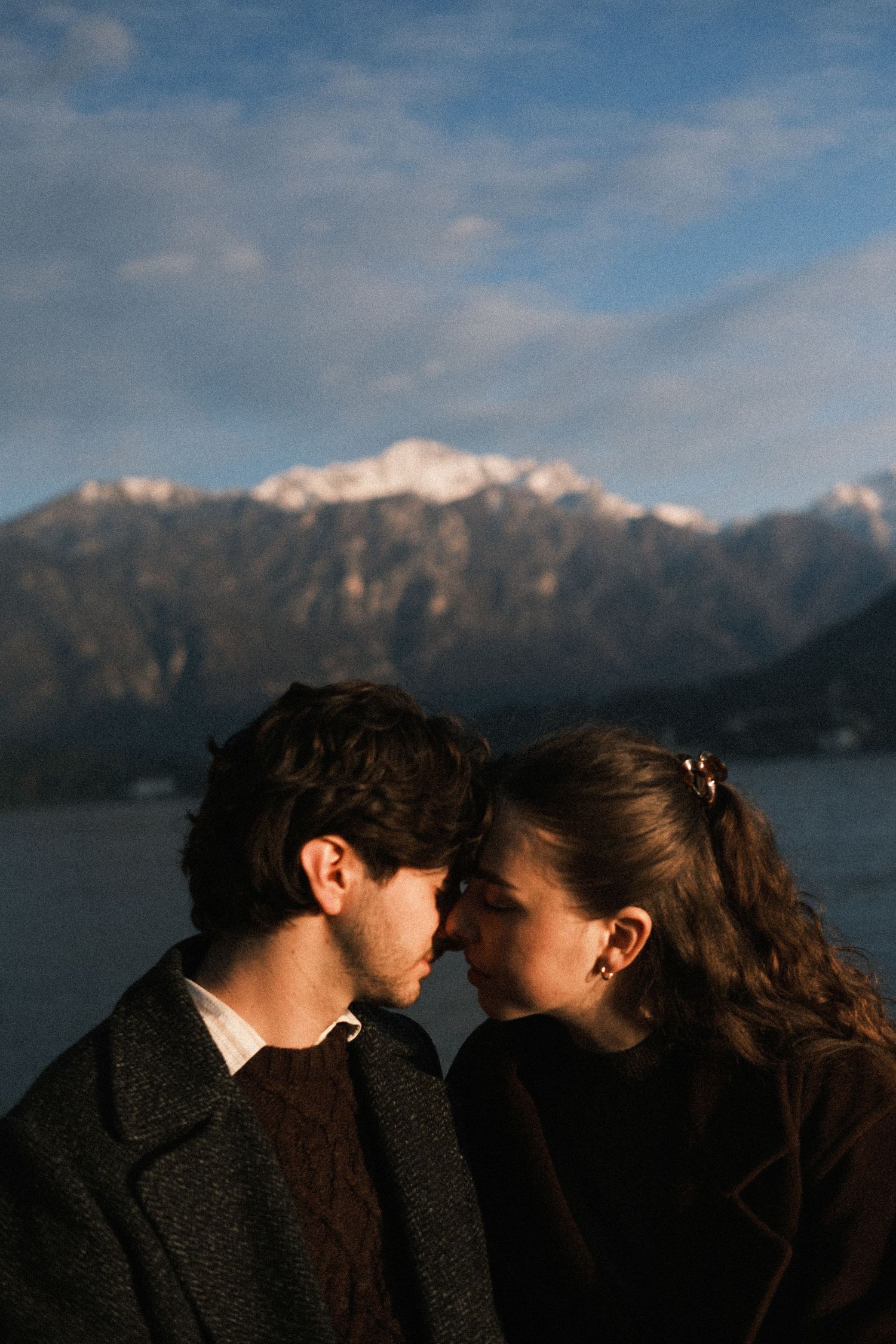 A couple in dark winter coats share an intimate moment against a backdrop of snow-capped mountains and a lake.
