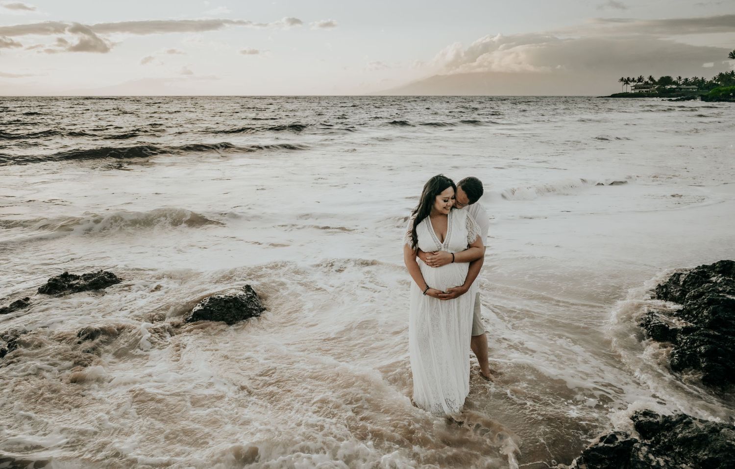Romantic beach maternity photo with waves crashing against rocks at sunset.