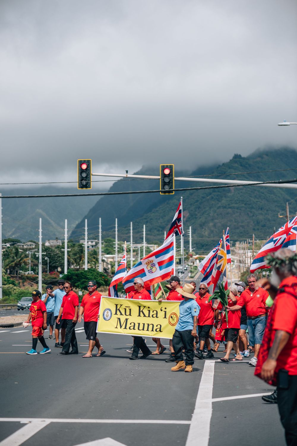 Protesters gather on a road with signs and Hawaiian flags while police vehicles block traffic.