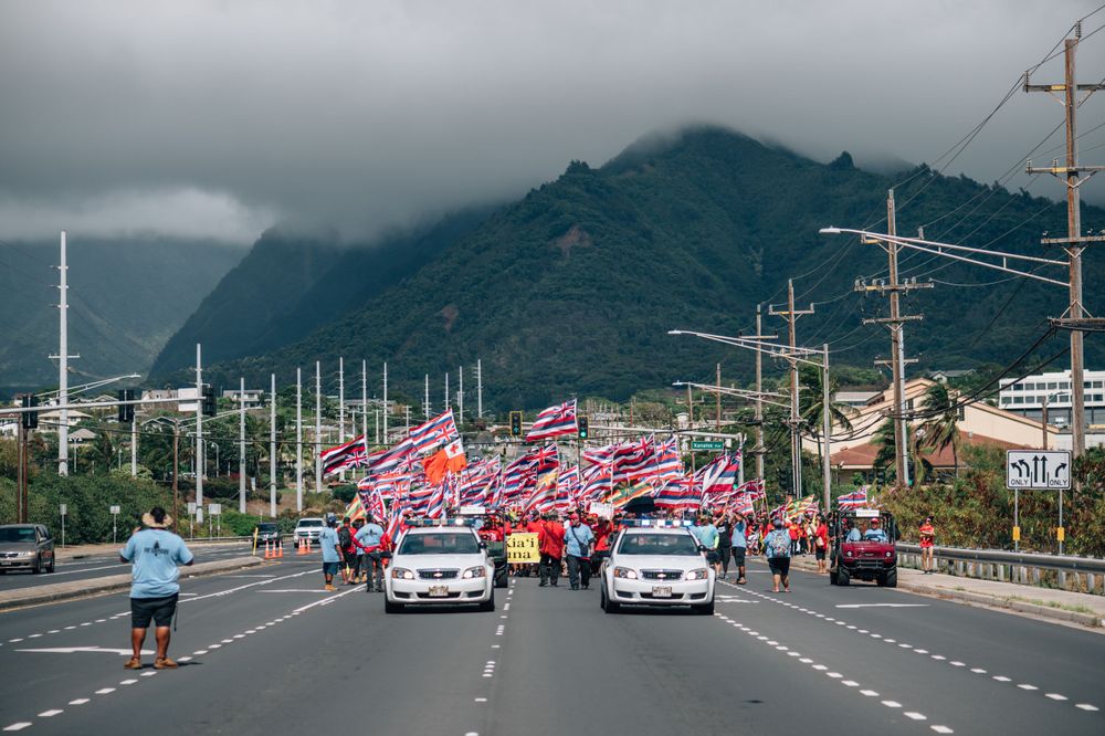 Large crowd with Hawaiian flags gathers along a road with mountains in the background.