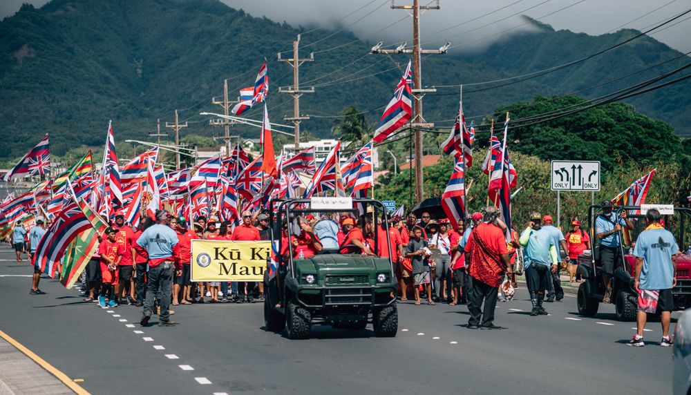 Procession of vehicles and flag-waving demonstrators march along a Hawaiian mountain road.