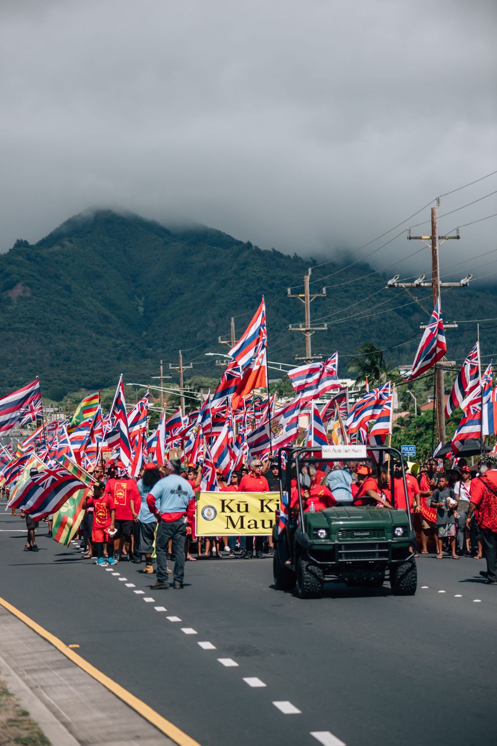 Rally participants wave Hawaiian flags while classic cars lead the procession on a mountain road.