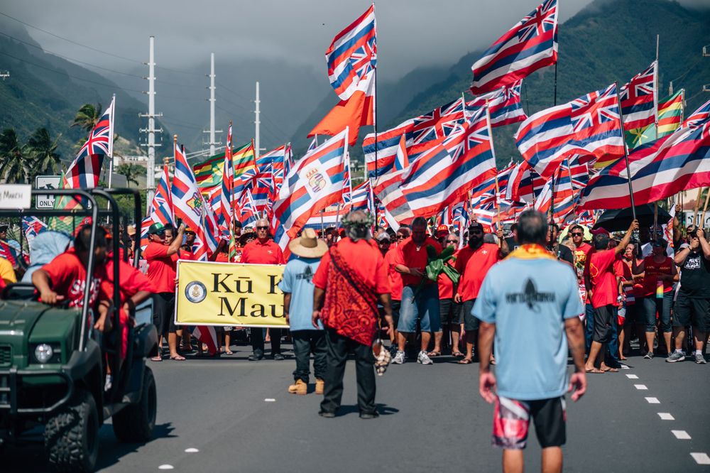 Demonstrators line a mountain road waving red and Hawaiian state flags during a protest.