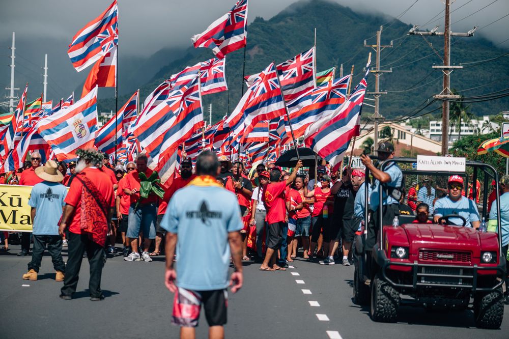 Crowd waves Hawaiian flags during protest march with mountains and cloudy skies in background.