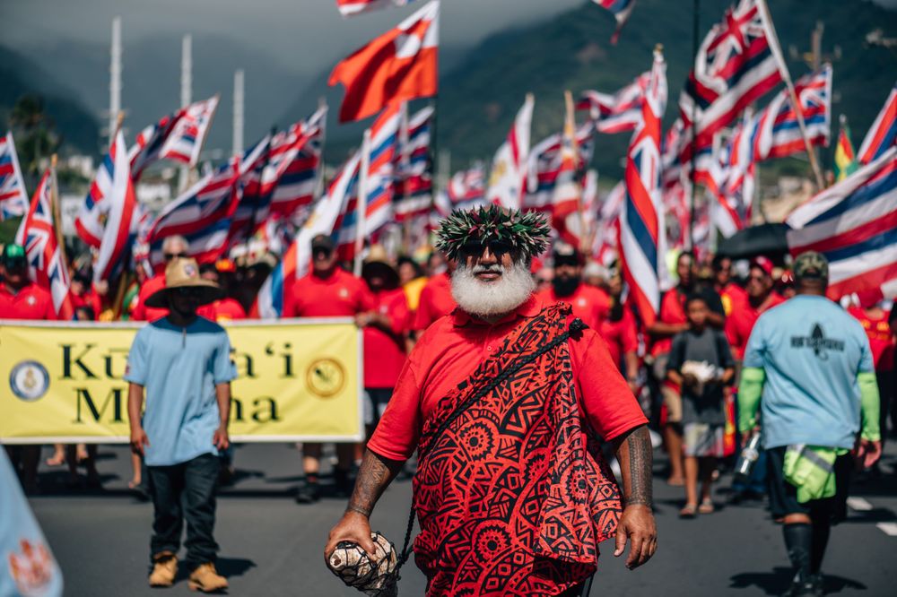 Large gathering of protesters with Hawaiian flags assemble along mountain roadway.