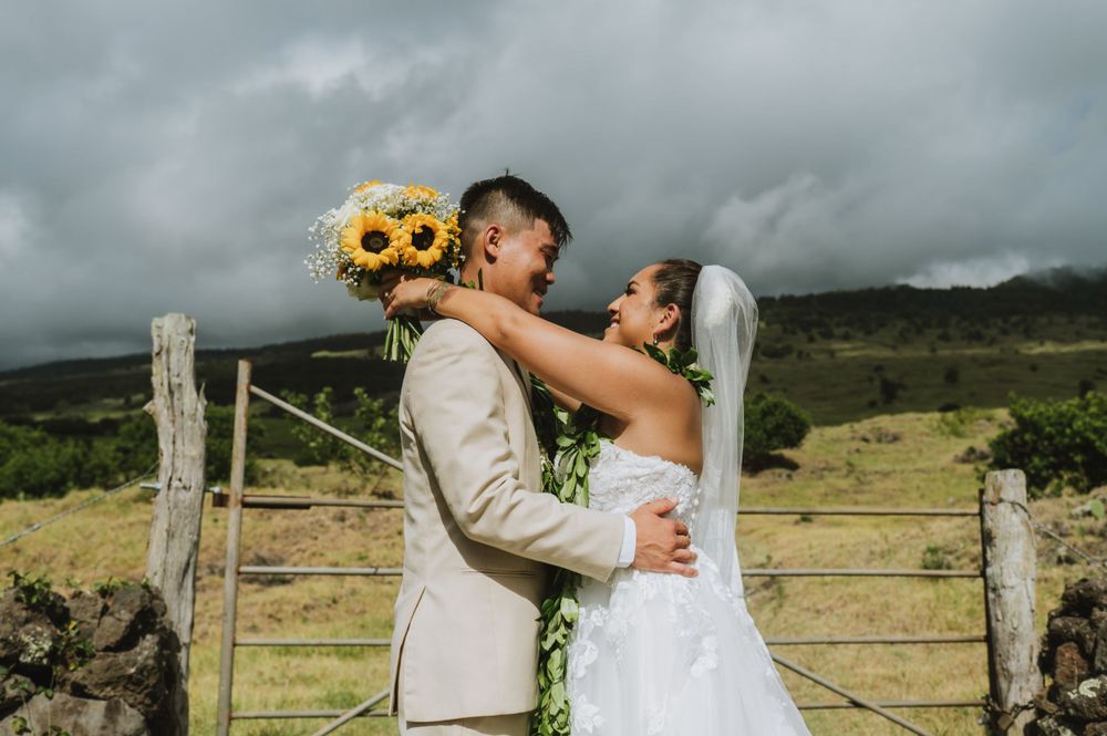 Newlyweds embrace against rustic fence with sunflowers and mountain landscape in background.