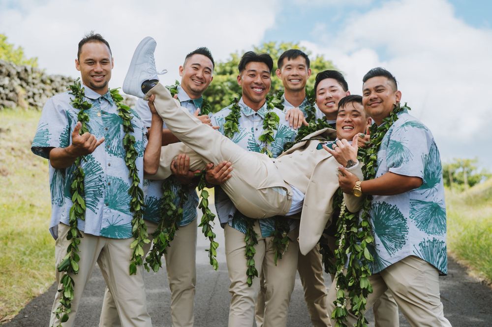 Groomsmen in tropical shirts playfully carrying groom in a light-colored suit outdoors.