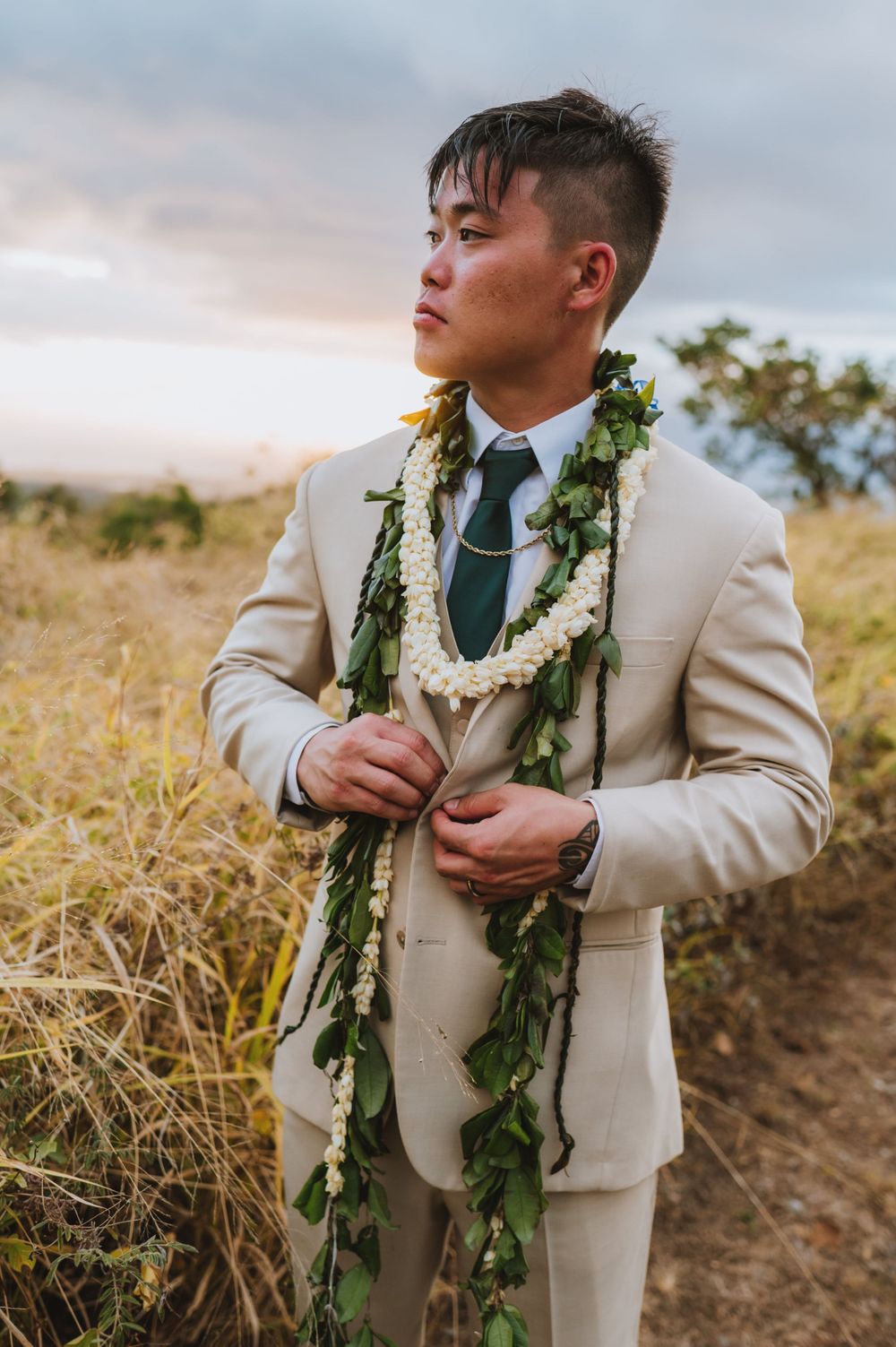 Groom in beige suit with traditional Hawaiian lei and greenery garland poses at sunset.