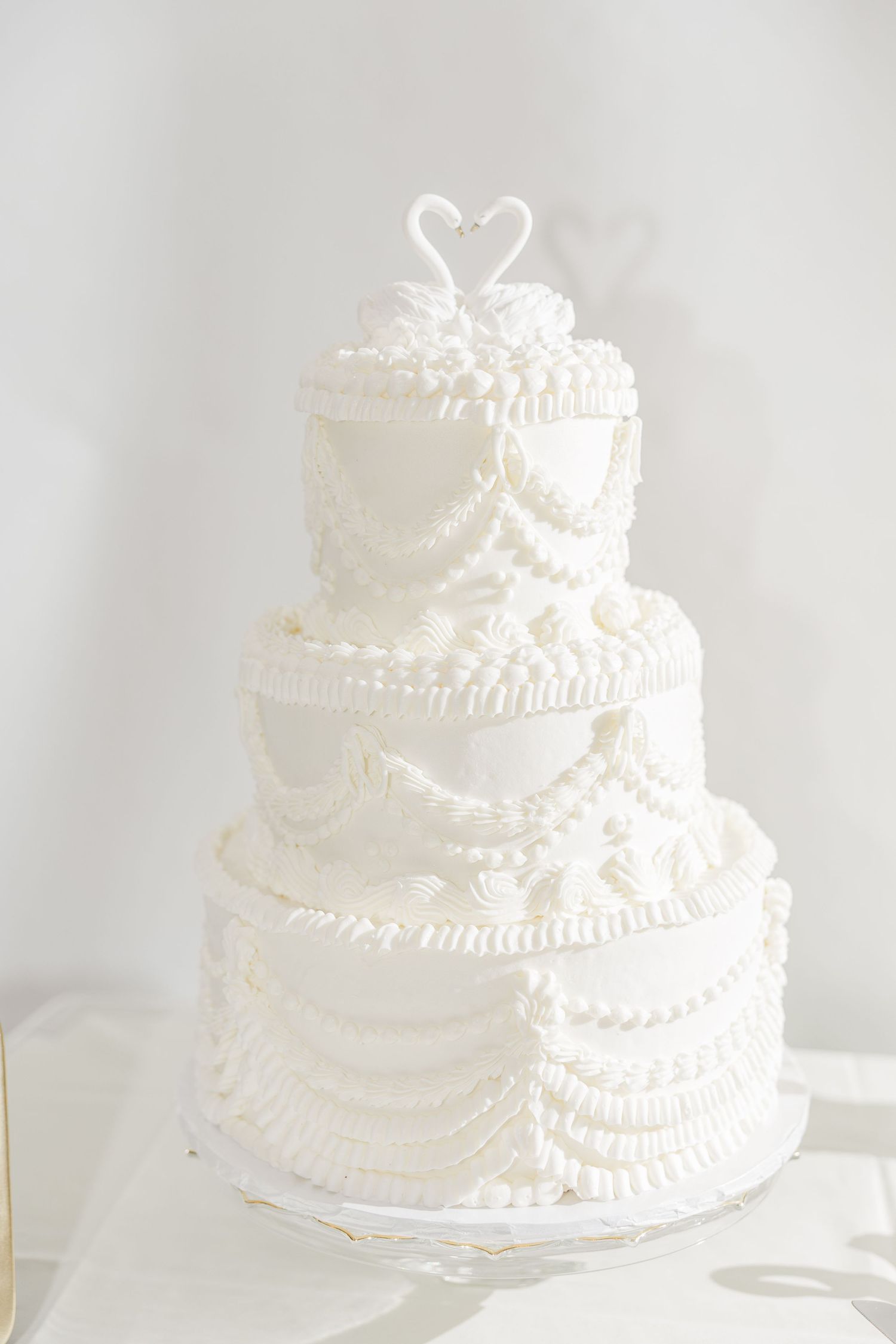 Elegant three-tiered white wedding cake with delicate piped lace details and pearl decorations against a light background.