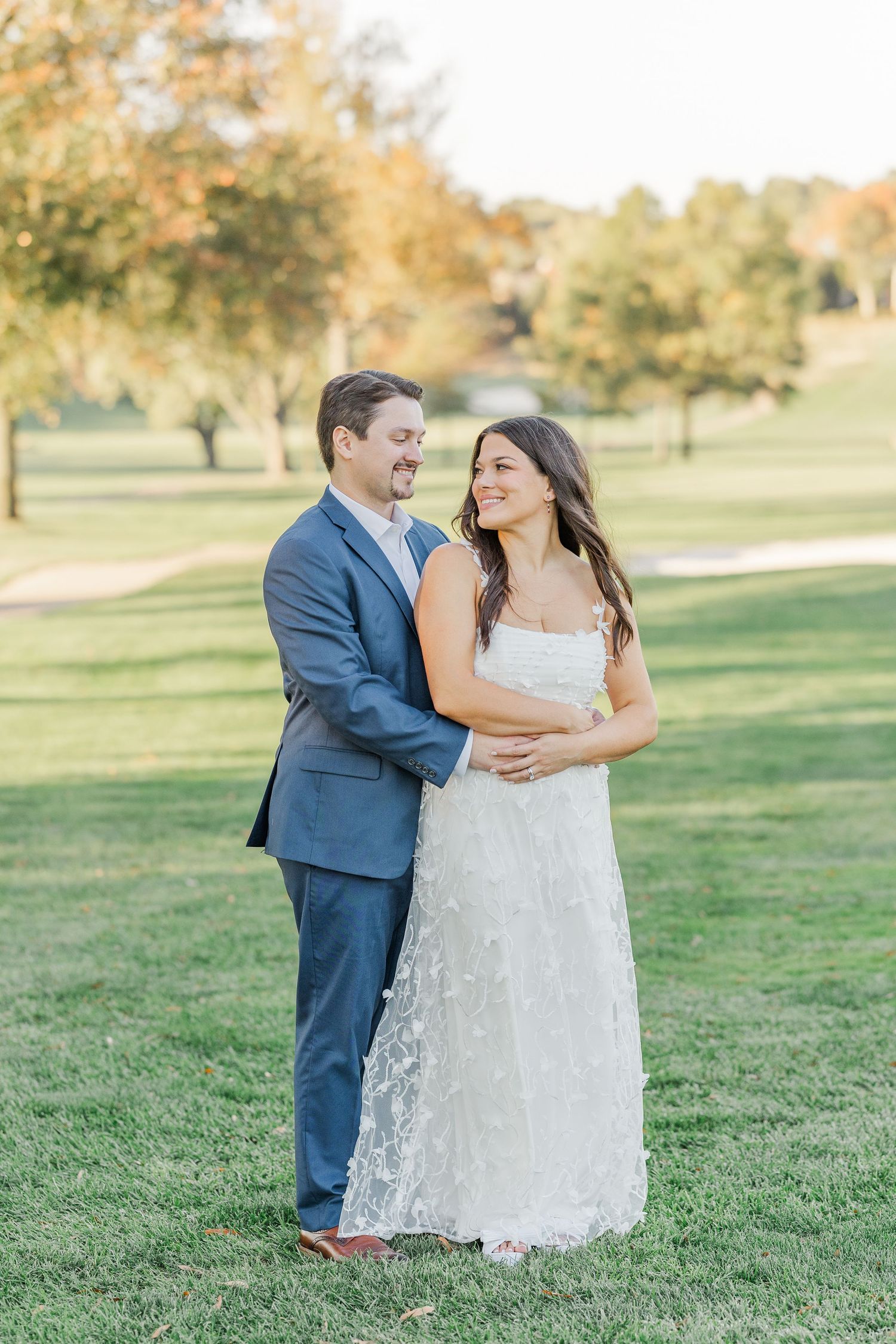 A couple in wedding attire shares a joyful moment together on a grassy lawn during an autumn day.