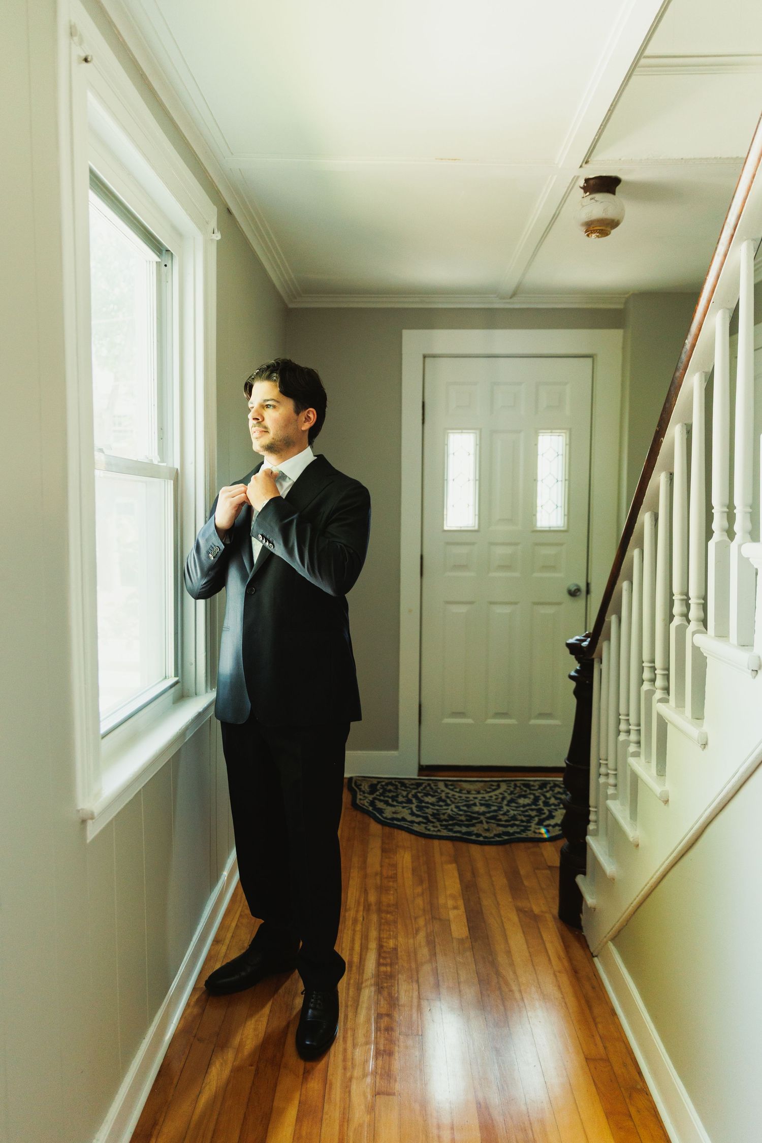 A formal suited figure adjusts their tie while standing in a sunlit hallway.