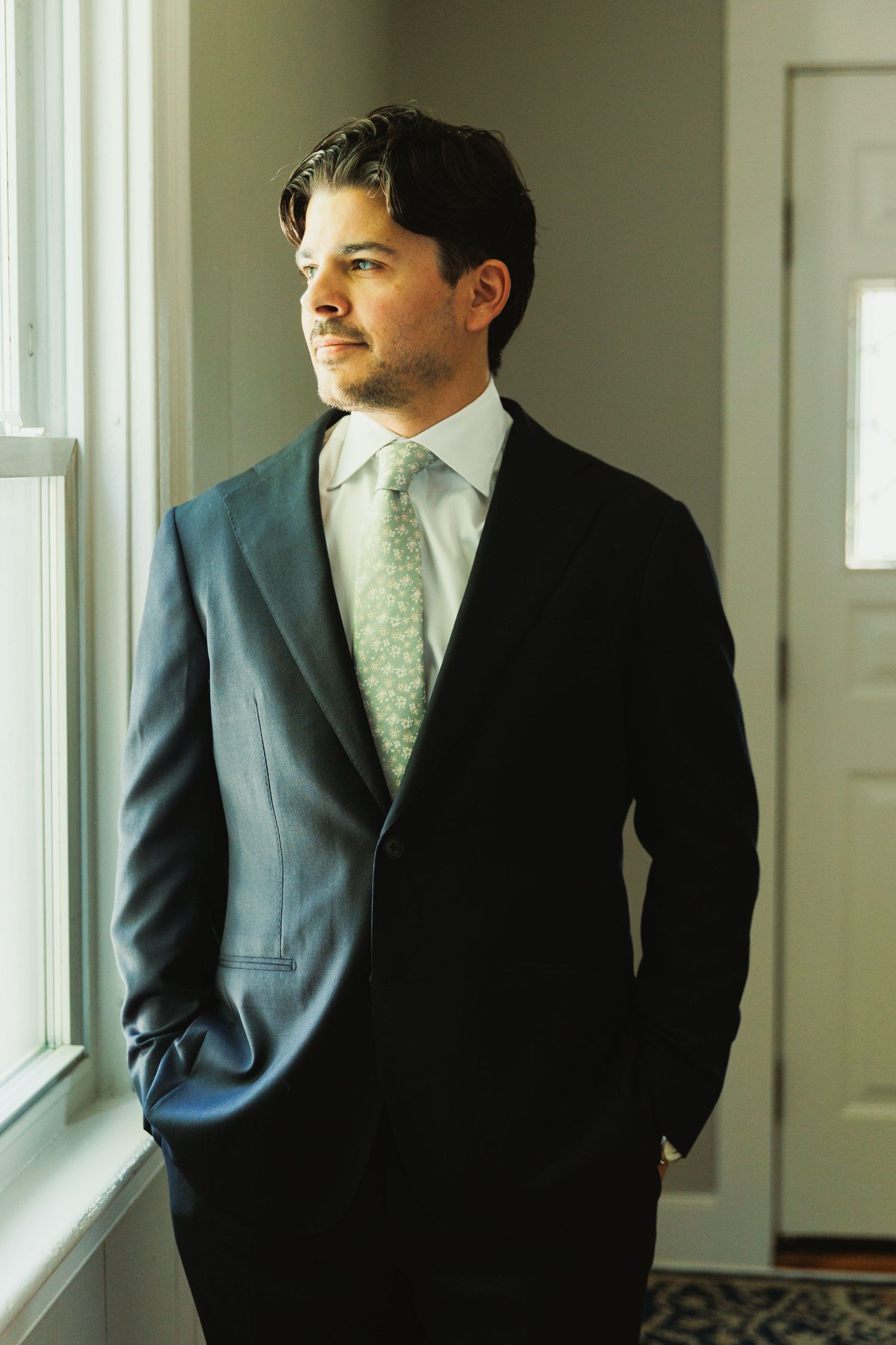 A suited figure gazes thoughtfully out a bright window in an elegant hallway.