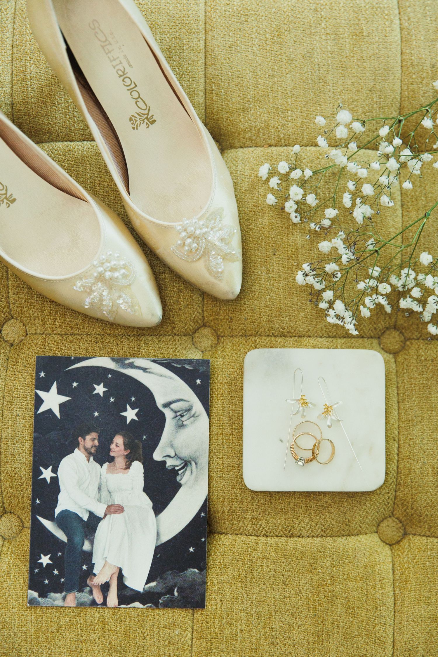 Wedding details flatlay with ivory shoes, baby's breath flowers, and wedding rings on yellow fabric.