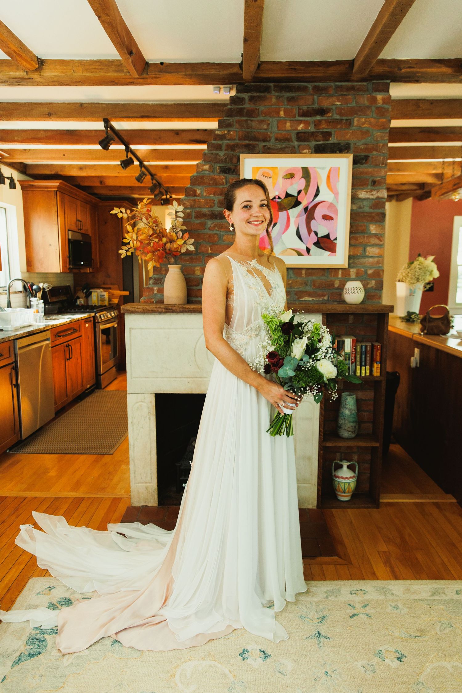 A person in a white wedding gown poses with flowers in an elegant room with exposed wooden beams.
