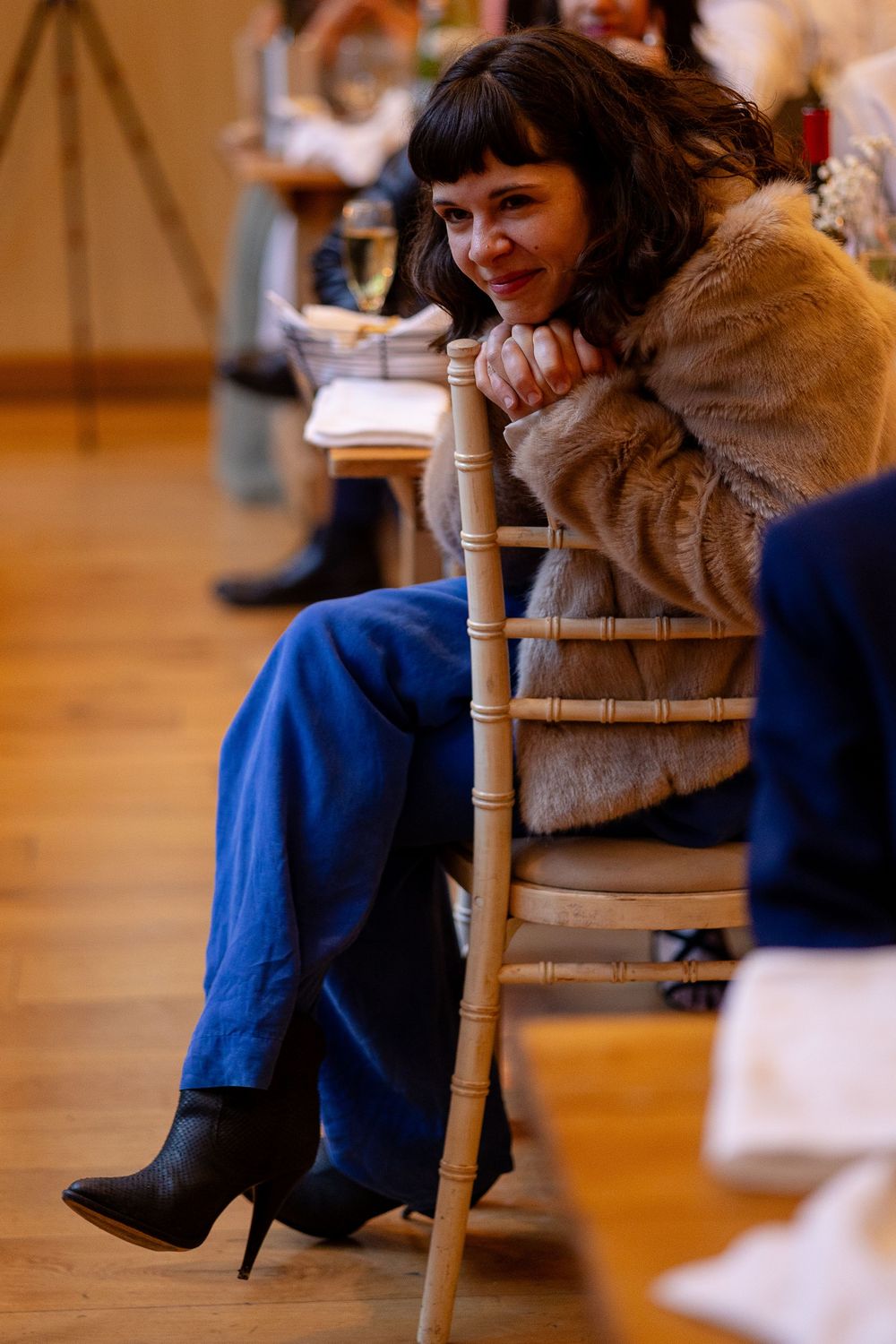 Close-up details of a wedding guest wearing a blue dress and fur stole while seated in a wooden chair.