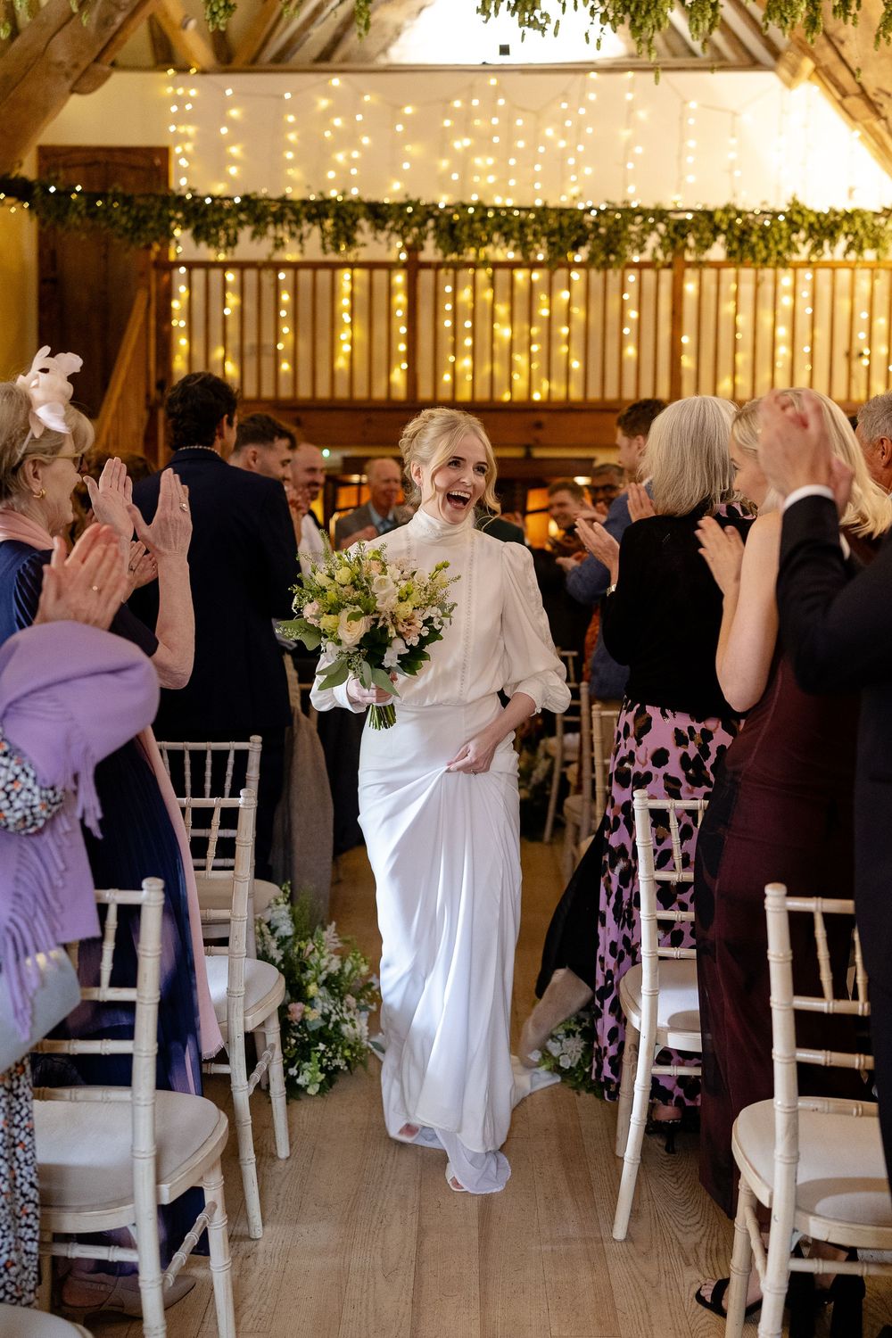 Wedding guests dancing and celebrating at an indoor reception with white chairs and string lights.